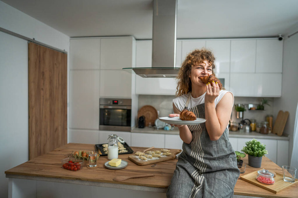 One adult caucasian woman stand in the kitchen with fresh croissant for breakfast daily morning routine - Photo, Image