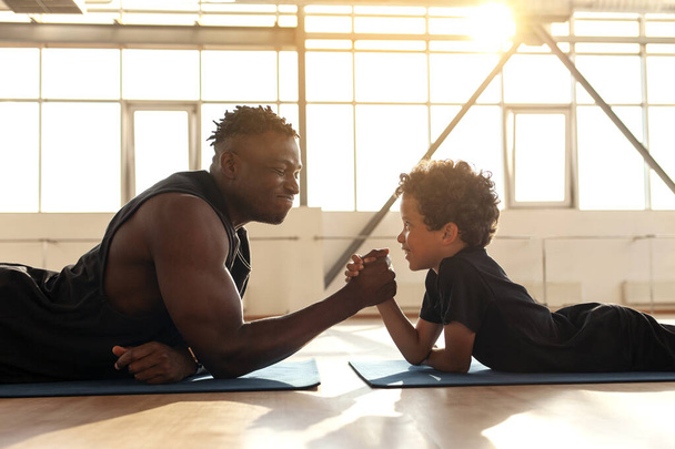 L'uomo afroamericano con suo figlio è impegnato nel braccio di ferro in palestra, papà misura la sua forza con suo figlio e insegna uno stile di vita sano - Foto, immagini