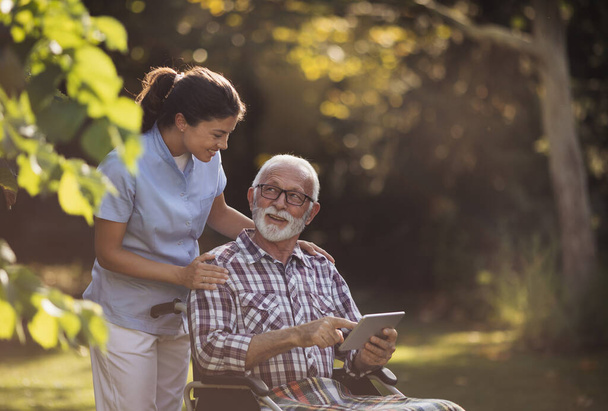 Hombre mayor en silla de ruedas mostrando algo en la tableta a la enfermera joven cuidador detrás de él en el parque - Foto, Imagen