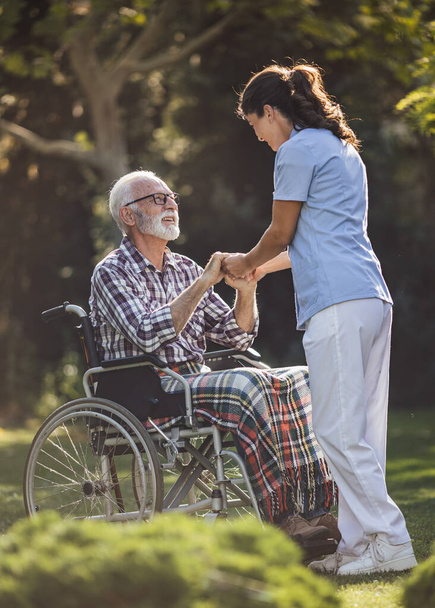 Νεαρή γυναίκα φροντιστής κρατώντας το χέρι του ηλικιωμένου άνδρα σε αναπηρική καρέκλα στο πάρκο - Φωτογραφία, εικόνα