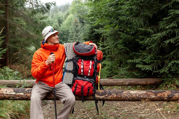 male tourist with backpack and hiking equipment sits resting in the forest, guy in orange jacket on mountain hike takes break - Photo, Image