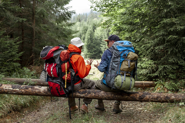 two male tourists with backpacks and hiking equipment sit in the forest and relax, people on a mountain hike talking during a break - Photo, Image
