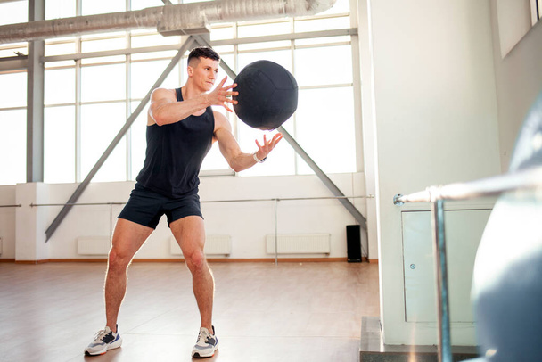 atletický mladý chlap ve sportovním oblečení na crossfit trénink dělá cvičení s míčem v jasné fitness místnosti, muž jde na sport a vlaky v tělocvičně - Fotografie, Obrázek