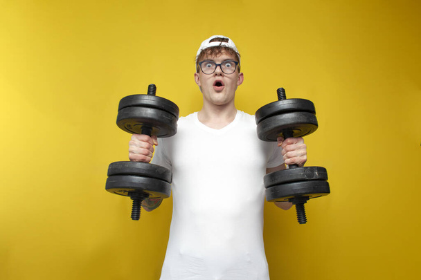 young thin, not athletic, nerd guy lifts heavy dumbbells and shows strength on a yellow background, a student goes in for sports with large weights - Foto, imagen