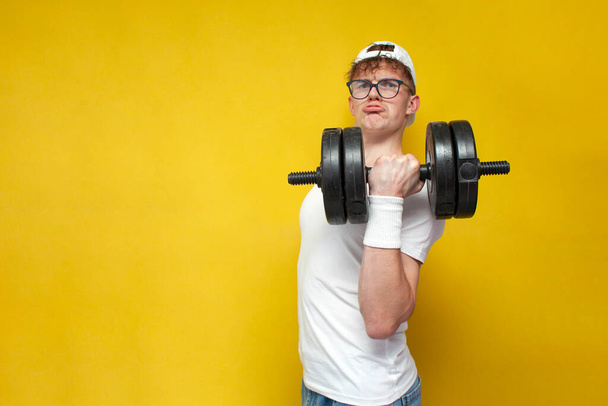 fiatal vékony, nem sportos, stréber srác szemüvegben emeli nehéz súlyzók és azt mutatja, erő sárga alapon, egy diák bemegy a sport nagy súllyal - Fotó, kép