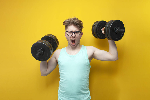 молодой уверенный парень студент поднимает тяжелые гантели на розовом фоне, мотивированный ботан в очках идет в спорт и кричит - Фото, изображение