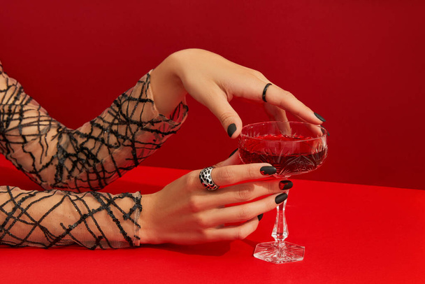 Γυναικεία χέρια με τρομακτικό σχεδιασμό νυχιών κρατώντας γυαλί με cranberry Halloween κοκτέιλ σε κόκκινο φόντο. Ματωμένο ποτό σε ρετρό ποτήρι. - Φωτογραφία, εικόνα