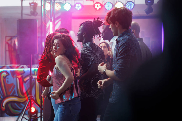 Νεαρή γυναίκα κάνει παθιασμένες χορευτικές κινήσεις ενώ κάνει clubbing με φίλους στη ντισκοτέκ. Πλήθος πάρτι σε ζωντανή πίστα ενώ φοιτούν ντίσκο πάρτι με ζωντανή μουσική σε νυχτερινό κέντρο διασκέδασης - Φωτογραφία, εικόνα