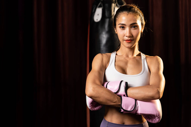 Ázsiai nő Muay Thai bokszoló puncs ököl előtt kamera készen áll a harcra állás pózol tornaterem boksz felszerelés a háttérben. Összpontosított szemek és felkészülés a kihívásra. Impetusz - Fotó, kép