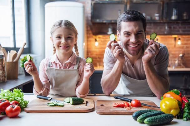 Cute little girl and her handsome dad are holding slices of cucumber and smiling while cooking in kitchen at home - Photo, Image