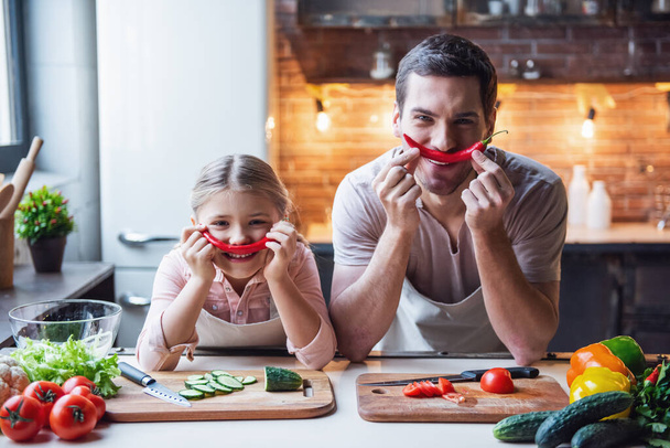 Sevimli küçük kız ve yakışıklı babası biber tutarak ve evde mutfakta yemek yaparken gülümseyen - Fotoğraf, Görsel