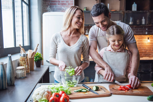 Linda niña y sus hermosos padres están cortando verduras y sonriendo mientras hacen ensalada en la cocina en casa
 - Foto, Imagen