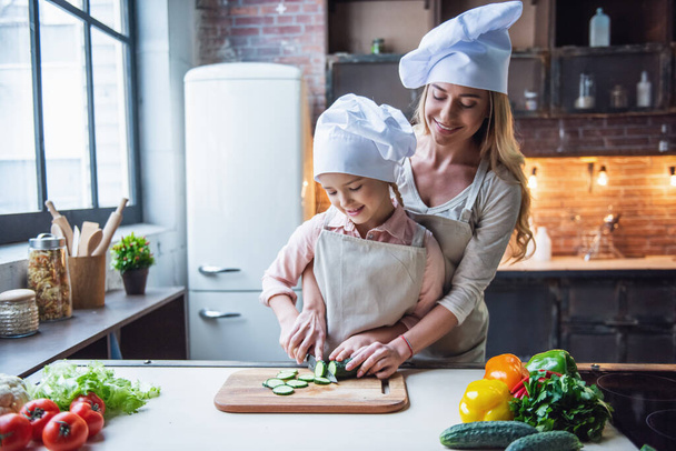 Menina bonito e sua bela mãe em chapéus do chef estão cortando legumes e sorrindo enquanto cozinha na cozinha em casa
 - Foto, Imagem