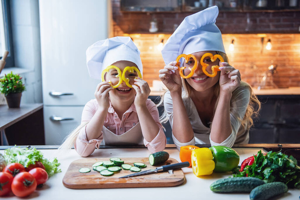 Cute little girl and her beautiful mom in chef's hats are having fun while cooking in kitchen at home - Photo, Image