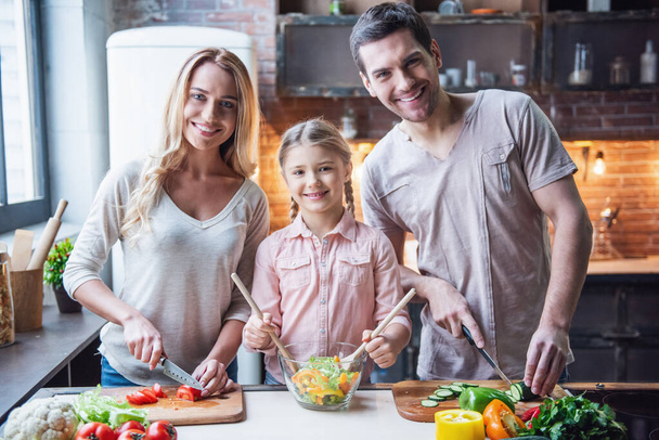 Cute little girl and her beautiful parents are cutting vegetables and smiling while making salad in kitchen at home - Photo, Image