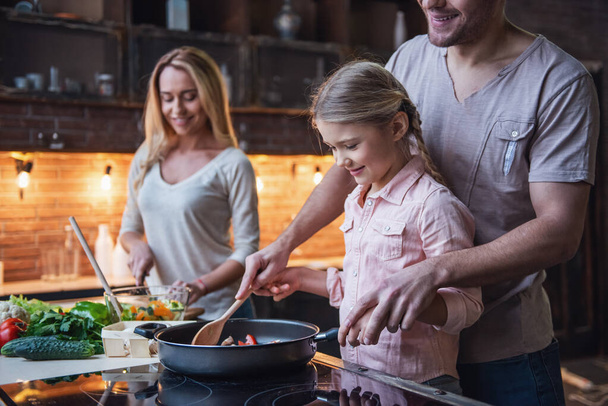 Cute little girl and her beautiful parents are  smiling while cooking in kitchen at home - Photo, Image