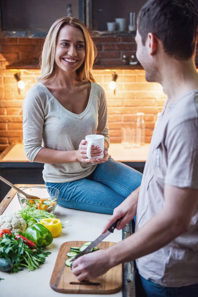 Hermosa mujer sostiene una taza y sonríe mientras su novio guapo está cocinando
 - Foto, Imagen