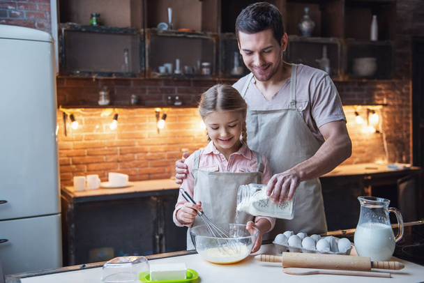 Linda niña y su padre guapo en delantales están batiendo huevos con harina y sonriendo mientras hornea en la cocina en casa
 - Foto, imagen