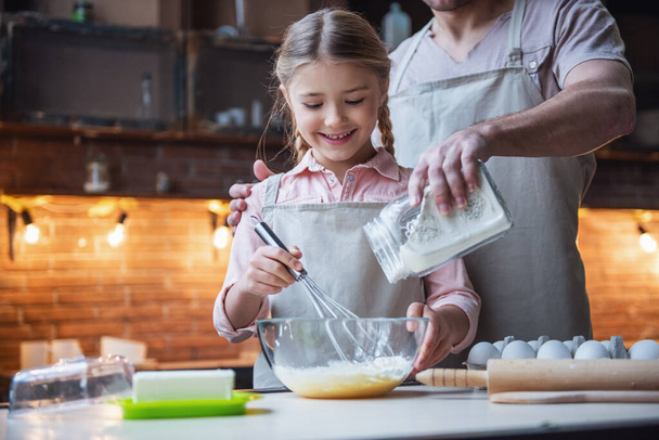 Το χαριτωμένο μικρό κορίτσι και όμορφος ο μπαμπάς της στις ποδιές χτυπώντας τα αυγά με το αλεύρι και χαμογελαστός κατά το ψήσιμο στην κουζίνα στο σπίτι - Φωτογραφία, εικόνα