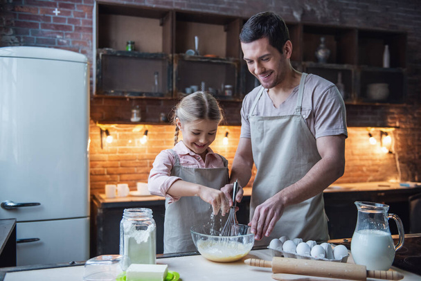 Nettes kleines Mädchen und ihr hübscher Vater in Schürzen verquirlen Eier und lächeln beim Backen in der heimischen Küche - Foto, Bild