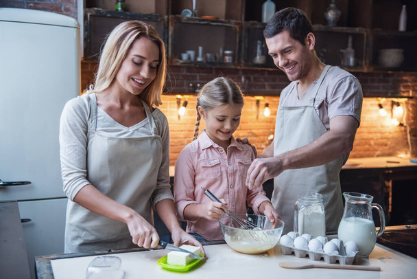 Nettes kleines Mädchen und ihre schönen Eltern in Schürzen lächeln beim Backen in der heimischen Küche - Foto, Bild