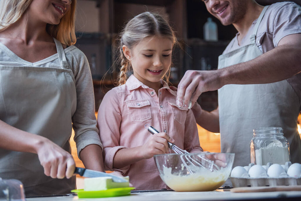 Το χαριτωμένο μικρό κορίτσι και όμορφη τους γονείς της σε ποδιές χαμογελαστός κατά το ψήσιμο στην κουζίνα στο σπίτι - Φωτογραφία, εικόνα