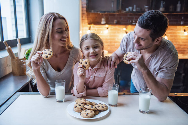 Carino bambina ei suoi bei genitori stanno mangiando biscotti con latte e sorridente mentre in piedi in cucina a casa
 - Foto, immagini