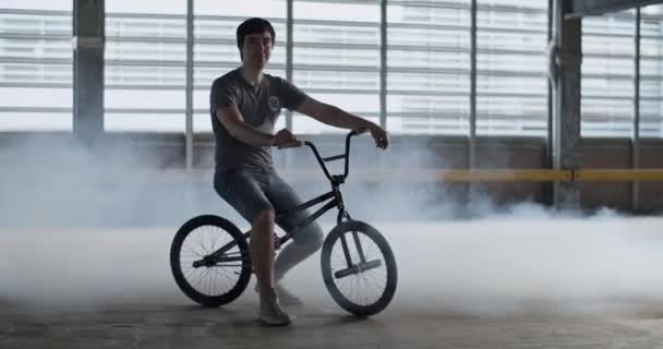 Full length handheld shot of young male rider in t shirt and shorts sitting on BMX putting hands on handlebar and foot on pedal inside spacious parking lot with smoke - Footage, Video