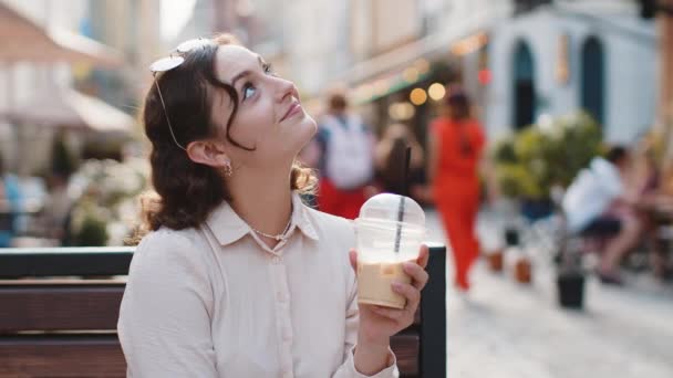 Boldog fiatal nő élvezi a reggeli hideg kávét jég és mosolygós szabadban. Pihentető, pihenő. A lány a padon ül a város napos utcájában, kávét iszik elvitelre. Városi életmód odakint - Felvétel, videó
