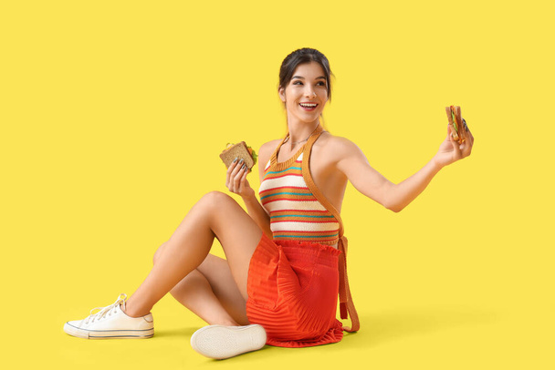 黄色の背景に座っているおいしいサンドイッチを持つ若い女性 - 写真・画像