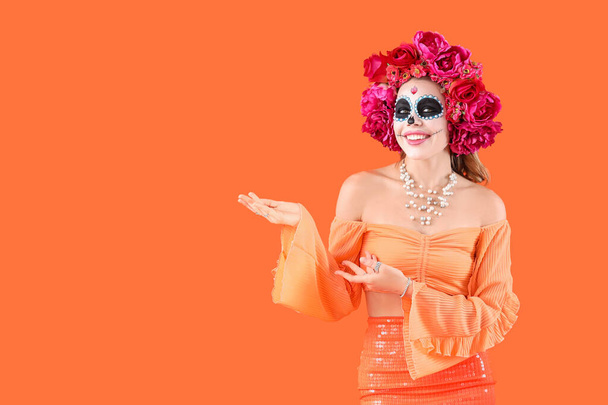 Mujer joven con el cráneo pintado mostrando algo sobre fondo naranja. Celebración del Día de los Muertos (El Día de Muertos) - Foto, Imagen