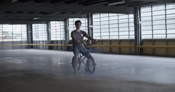 Ganzkörperaufnahme eines jungen Sportlers auf BMX sitzend mit der Hand am Griff während des Trainings auf einem großzügigen Parkplatz und Blick in die Kamera - Filmmaterial, Video