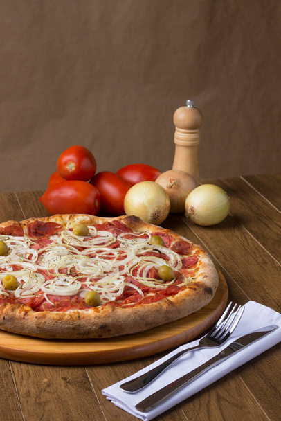 Foto gastronomica di pizza al salame piccante con fette di cipolla, mozzarella e olive verdi. Ingredienti nella loro forma rustica per comporre l'immagine. - Foto, immagini