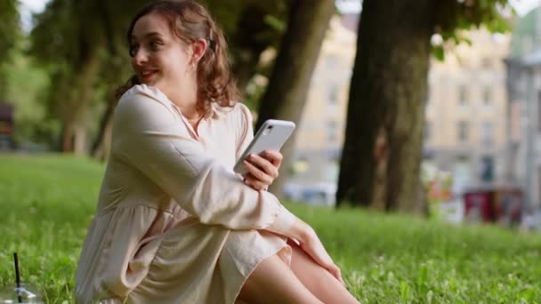 スマートフォンの入力テキストメッセージを使用して笑顔の若い女性は、モバイル画面のソーシャルメディアアプリを見てオンラインチャットメッセージに応答します。都市部の夕焼け都市公園で草の上に座っている女の子手に電話を保持 - 映像、動画