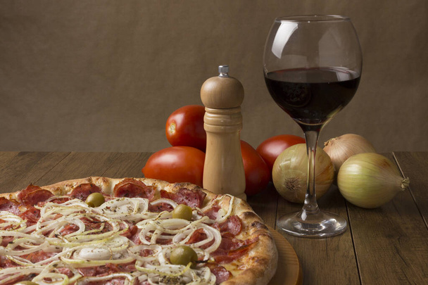 Pepperoni pizza met plakjes uien, mozzarella kaas en groene olijven. Glas wijn, uien en rustieke tomaten. Horizontale Gastronomische Fotografie met linksboven ruimte voor teksten. - Foto, afbeelding