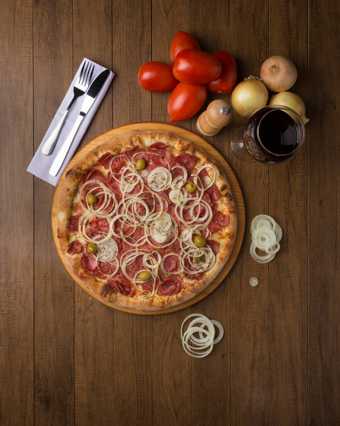 Pepperoni pizza met plakjes uien en groene olijven. Glas rode wijn, uien en rustieke tomaten. Top gastronomische fotografie. - Foto, afbeelding