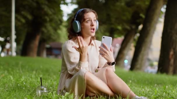 ワイヤレスヘッドフォンを選択し、スマートフォンのダンス屋外で好きな精力的なディスコロックnロール音楽を聞いて、幸せなリラックスした喜びの若い女性。都会の夕焼け都市公園で草の上に座っている女の子 - 映像、動画