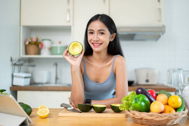 Portret atrakcyjnej i uśmiechniętej Azjatki w stroju gimnastycznym pokazuje awokado przed kamerą podczas przygotowywania zdrowych posiłków w kuchni. - Zdjęcie, obraz