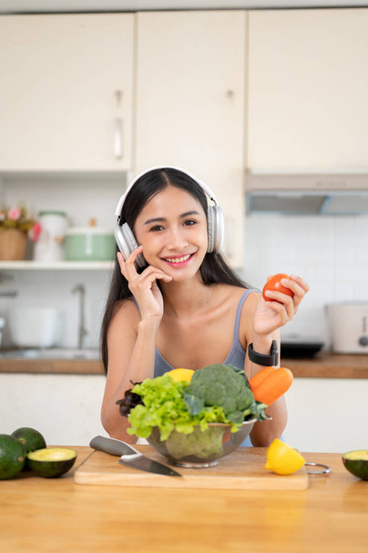 Ein Porträt einer hübschen Asiatin in Turnbekleidung lächelt in die Kamera und genießt die Musik auf ihrem Kopfhörer, während sie in der Küche Zutaten für ihr gesundes Frühstück und Smoothie zubereitet.. - Foto, Bild