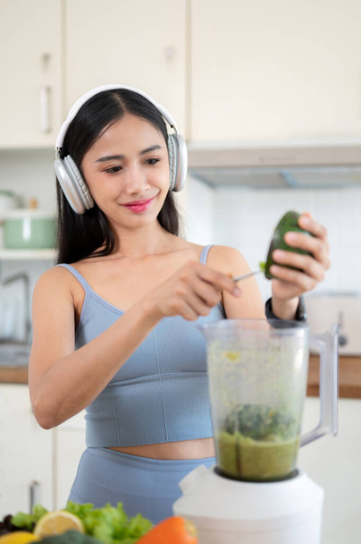 Egy gyönyörű és karcsú, fiatal ázsiai nő tornaruhában és fejhallgatóban avokádót ad egy turmixgéphez, így egészséges zöld turmixlevet készít a konyhában reggeli edzés után.. - Fotó, kép