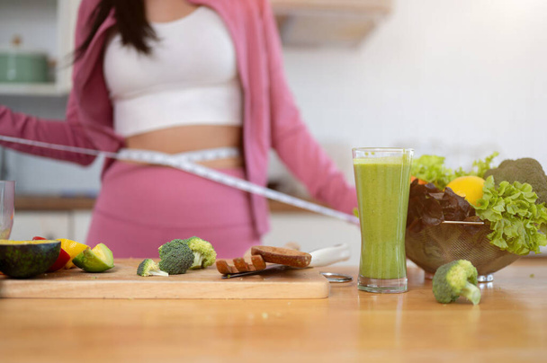 Close-up image of a glass of a healthy green smoothie and fresh vegetables on a kitchen table in the kitchen with a female measuring her waist with a measuring tape in the background. Diet recipes - Photo, Image