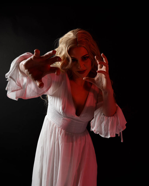 Κοντινό πορτρέτο της τρομακτικής ξανθιάς νύφης βαμπίρ φορώντας κομψό λευκό αποκριάτικο φόρεμα φαντασίας. Απομονωμένα σε σκοτεινό φόντο στούντιο με αγχώδη χέρια gestural φτάσει έξω  - Φωτογραφία, εικόνα