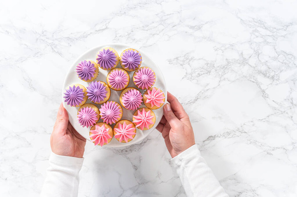 Flat lay. Freshly baked mini vanilla cupcakes with ombre pink buttercream frosting on a white cake stand. - Photo, Image