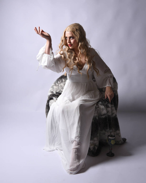 Retrato de cuerpo entero de mujer rubia vestida con vestido de novia histórico blanco vestido de fantasía. Posar sentado con las manos gestuales extendiéndose, aislado en el fondo del estudio. - Foto, Imagen