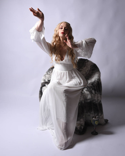Pełna długość portret blondynki noszącej białą historyczną suknię ślubną strój fantasy. Siedząca poza z gestywnymi dłońmi wyciągniętymi do góry, odizolowana na tle studia. - Zdjęcie, obraz