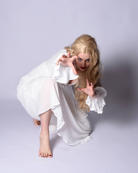 Pełna długość portret blondynki noszącej białą historyczną suknię ślubną strój fantasy. Siedzenie na podłodze z gestywnymi dłońmi wyciągniętymi do góry, odizolowane na tle studia. - Zdjęcie, obraz