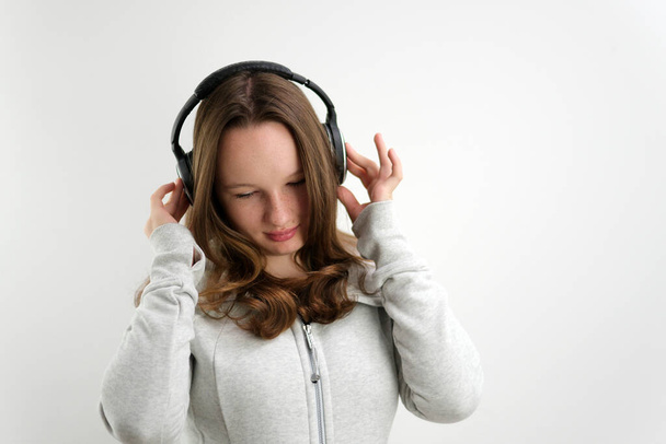 έφηβος κορίτσι ακούει μουσική σε μεγάλα επαγγελματικά ακουστικά σε λευκό φόντο αθλητικό κοστούμι χαμογελά απαλά όμορφη έφηβη. εφηβεία - Φωτογραφία, εικόνα