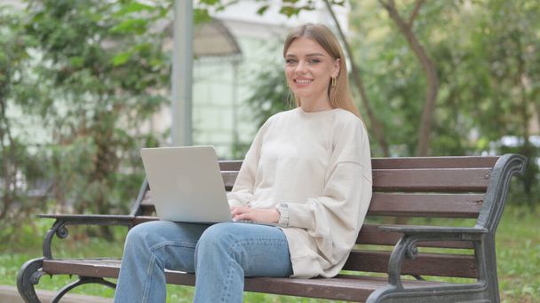Lässige junge Frau lächelt in die Kamera, während sie draußen am Laptop arbeitet - Foto, Bild