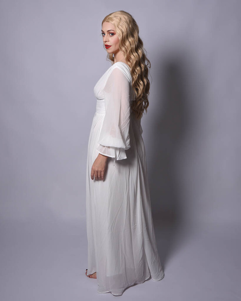 Pełna długość portret blondynki noszącej białą historyczną suknię ślubną strój fantasy. Pozycja stojąca, zwrócona tyłem do kamery, odizolowana na tle studia. - Zdjęcie, obraz