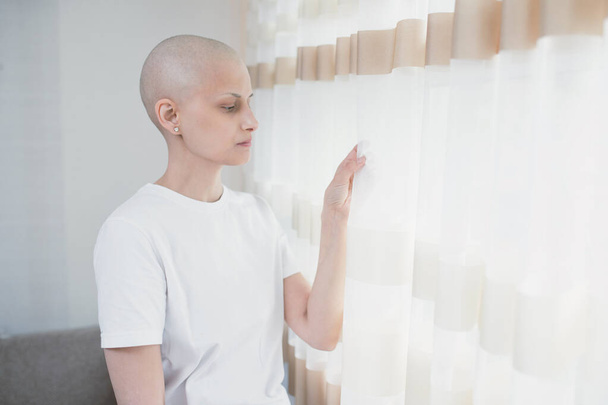 Νεαρή άρρωστη φαλακρή γυναίκα ασθενής με ογκολογική πάλη με καρκίνο κοιτάζει το παράθυρο, αισθάνεται μοναξιά στο σπίτι, ελπίδα για μελλοντική ανάρρωση. - Φωτογραφία, εικόνα